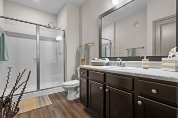 A bathroom with hardwood floors, dark cabinets and drawers, a vanity sink with a large mirror and great lighting, and a walk-in shower with sliding glass doors.